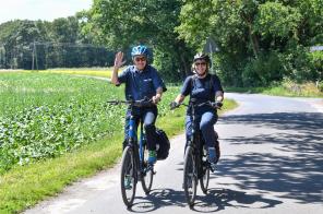 Zwei Radfahrer fahren der Kamera entgegen und winken